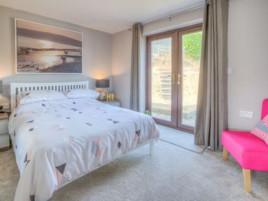 A bedroom with a bed bedside tables lamps a pink chair and glass doors leading outside at Cromlin Glas in Abersoch