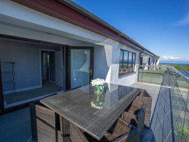 An outdoor balcony with a glass-top dining table and wicker chairs overlooking the sea at Cromlin Glas in Abersoch