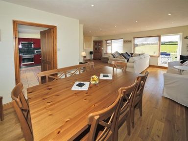 Dining area with wooden table and chairs next to a living room with sofas and large windows at Eleri in Abersoch