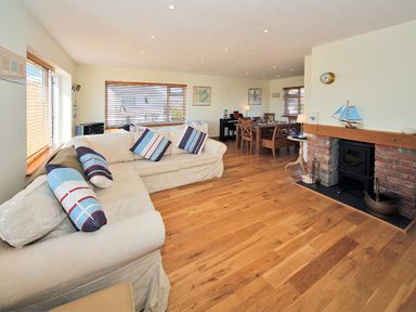 A living room with a beige sectional sofa and striped cushions near windows a dining table and chairs a fireplace with a wood stove and wooden floor at Eleri in Abersoch