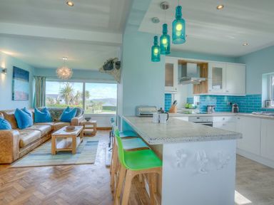 A living room with a brown sofa and blue pillows next to a kitchen with a white counter and green and blue chairs at Gilfach in Abersoch