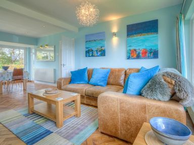 A living room with a brown leather sofa and blue cushions next to a wooden coffee table and dining area in the background at Gilfach in Abersoch