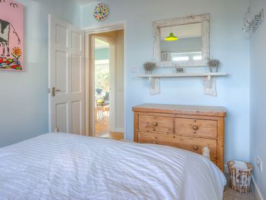 A bedroom with a white bedspread a wooden chest of drawers a white framed mirror and an open door showing a dining area at Gilfach in Abersoch