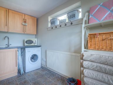 A utility room with a washing machine microwave sink wooden cabinets and stacked cushions at Gilfach in Abersoch