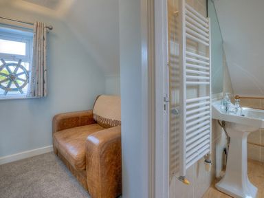 A small room with a brown sofa and a window with a ship wheel curtain tieback next to a bathroom with a white towel radiator and pedestal sink at Gilfach in Abersoch