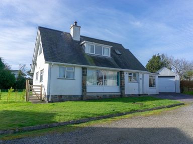 A detached house with a sloped roof and chimney on a grass lawn with a gravel driveway and a garage at Gilfach in Abersoch
