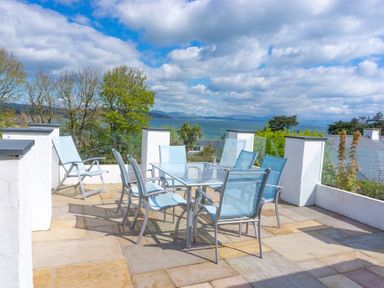A patio with a glass table and chairs overlooking trees and water at 3 Madoc View in Bwlchtocyn near Abersoch