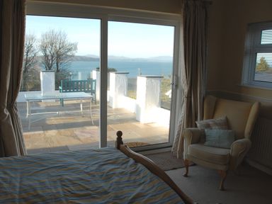 A bedroom with a bed and an armchair next to sliding glass doors leading to a patio with a table and bench overlooking the sea at 3 Madoc View in Bwlchtocyn near Abersoch