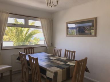 A dining room with a table covered by a checkered tablecloth and wooden chairs near a window with curtains at 3 Madoc View Bwlchtocyn near Abersoch