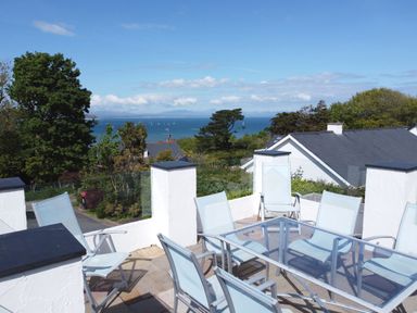 An outdoor patio with glass table and blue chairs overlooking trees rooftops and the sea at 3 Madoc View Bwlchtocyn near Abersoch