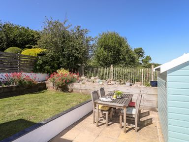 A garden with a wooden dining table and chairs on a patio next to a grass lawn and a light blue shed at The Anchorage in Abersoch