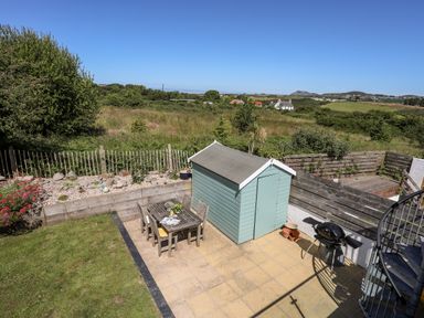 An outdoor patio with a wooden table and chairs a small shed a grill and a spiral staircase overlooking a grassy field at The Anchorage in Abersoch