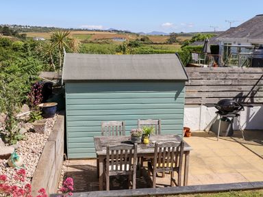A garden with a wooden table and chairs next to a green shed and barbecue at The Anchorage in Abersoch