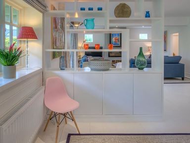 A white shelf divider with decorative glass items and a pink chair next to a window with a plant and lamp at The Coach House in Abersoch