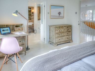 A bedroom with a wooden desk with a laptop and a pink chair a wooden chest of drawers and a bed with a grey blanket at The Coach House in Abersoch