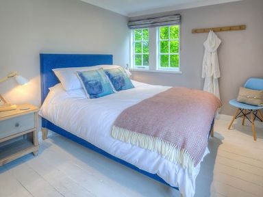 A bedroom with a blue bed frame white bedding blue pillows a pink blanket wooden nightstand with a lamp and a blue chair with a cushion at The Coach House in Abersoch