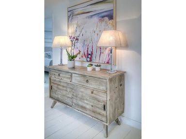 A wooden sideboard with two table lamps and three small potted plants with a large abstract painting above at The Coach House in Abersoch