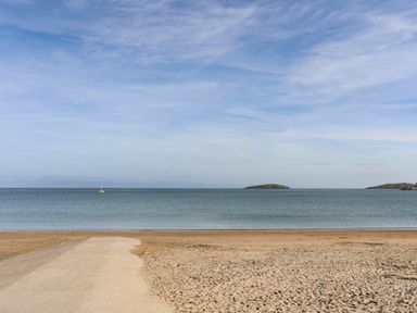 A sandy beach with a concrete path leading to the sea and a yellow sailboat on the water at The Warren D26 in Abersoch