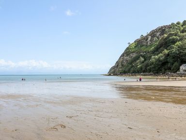 A sandy beach with people walking and a rocky hillside with trees at The Warren D26 in Abersoch