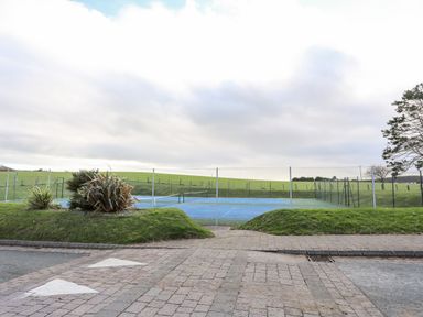 A tennis court with net and blue surface behind a fence with grass and plants in the foreground at The Warren D26 in Abersoch
