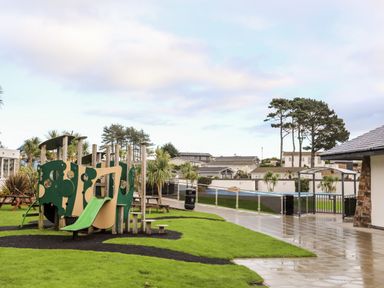 An outdoor playground with a slide and climbing structures near a paved walkway and picnic tables at The Warren D26 in Abersoch
