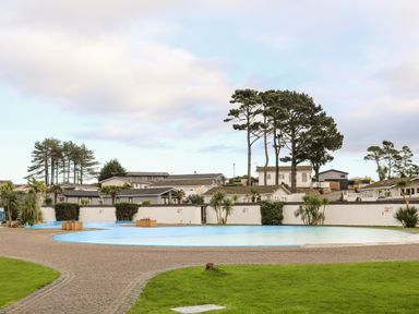 An outdoor pool with surrounding pavement and grass with houses and trees in the background at The Warren D26 in Abersoch