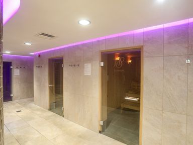 A hallway with tiled walls and floors leading to two wooden door saunas with glass fronts and purple lighting on the ceiling at The Warren D26 in Abersoch