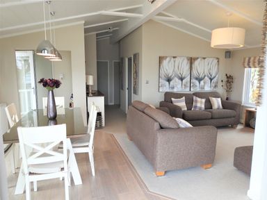 A dining area with a glass table and white chairs next to a living room with gray sofas and paintings on the wall at The Warren D26 in Abersoch