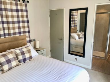 A bedroom with a bed with checkered pillows and curtains a nightstand with a lamp a door a full length mirror and an entrance to a tiled shower at The Warren D26 Abersoch