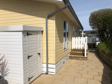 A paved outdoor area with a yellow building, white storage shed, wooden steps, white railing, and some greenery at The Warren D26 in Abersoch