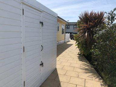 A paved walkway between a white shed and plants with a yellow building and a car in the background at The Warren D26 in Abersoch