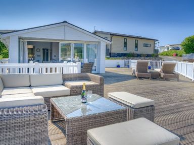 An outdoor patio with wicker seating, a glass table, and a bottle with glasses at The Warren Q4 in Abersoch