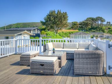 An outdoor patio area with wicker sofa set and table on wooden deck with houses and trees in the background at The Warren Q4 in Abersoch