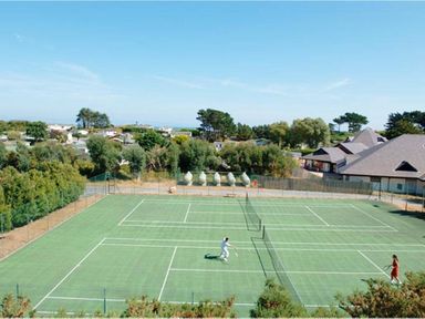 An outdoor tennis court with two people playing surrounded by trees and buildings at The Warren T23 in Abersoch