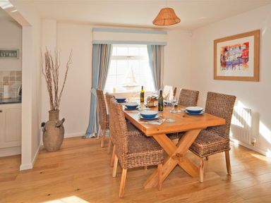 A dining room with a wooden table set for six wicker chairs and a window with blue curtains at Ty Coed Abersoch