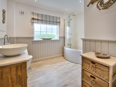 A bathroom with a bathtub and shower a wooden cabinet with drawers a sink with a faucet and a frosted window at Ty Coed  in Abersoch