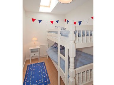 A small bedroom with white bunk beds a blue rug a side table with a lamp and a skylight at Ty Coed in Abersoch