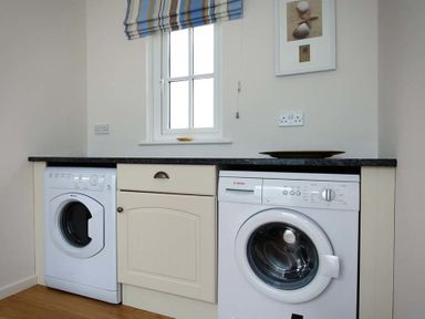 A laundry room with washing machines under a countertop beneath a window at Ty Coed in Abersoch