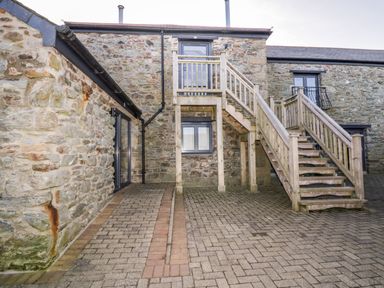 An outdoor stone courtyard with wooden stairs leading to an upper door at Beacon Barn in St Agnes