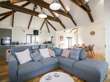An open plan living area with a grey sectional sofa with cushions a round coffee table a dining table with chairs and a kitchen with bar stools and visible ceiling beams at Beacon Barn in St Agnes