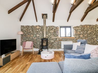 A living room with stone walls wooden beams a wood stove a grey sofa and a pink armchair at Beacon Barn in St Agnes