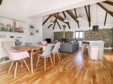 An open living room with a dining table and chairs a gray sofa a kitchen island with stools wooden beams and stone walls at Beacon Barn in St Agnes