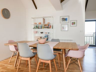 A dining room with a wooden table, six chairs, a white bookshelf with books and bowls, a wall clock, and framed pictures at Beacon Barn in St Agnes