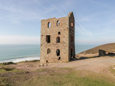A stone building ruin on a grassy and rocky coastal cliff with ocean in the background at Beacon Barn in St Agnes