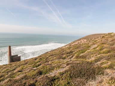 A hillside covered with shrubs overlooking the ocean with a tall chimney structure near the shore at Beacon Barn in St Agnes