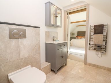 A bathroom with a toilet sink cabinet mirror cabinet and towel radiator with a bedroom visible through the door at Stow Cottage Barn in Churchill Oxfordshire