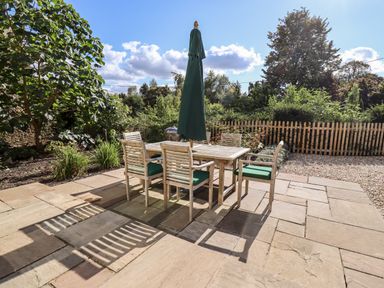 An outdoor patio with a wooden table and six chairs with green cushions and a closed green umbrella surrounded by plants and a wooden fence at Stow Cottage Barn in Churchill Oxfordshire