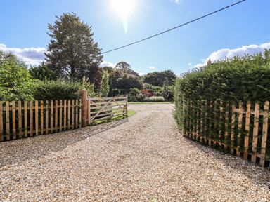 A gravel driveway with wooden gates and fencing surrounded by hedges and trees at Stow Cottage Barn in Churchill Oxfordshire