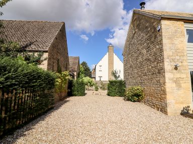 An outdoor space with gravel ground between two stone buildings with bushes and green plants at Stow Cottage Barn in Churchill Oxfordshire