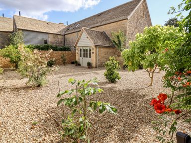 An outdoor courtyard with gravel ground various small trees and plants and a stone building with a white door at Stow Cottage Barn in Churchill Oxfordshire
